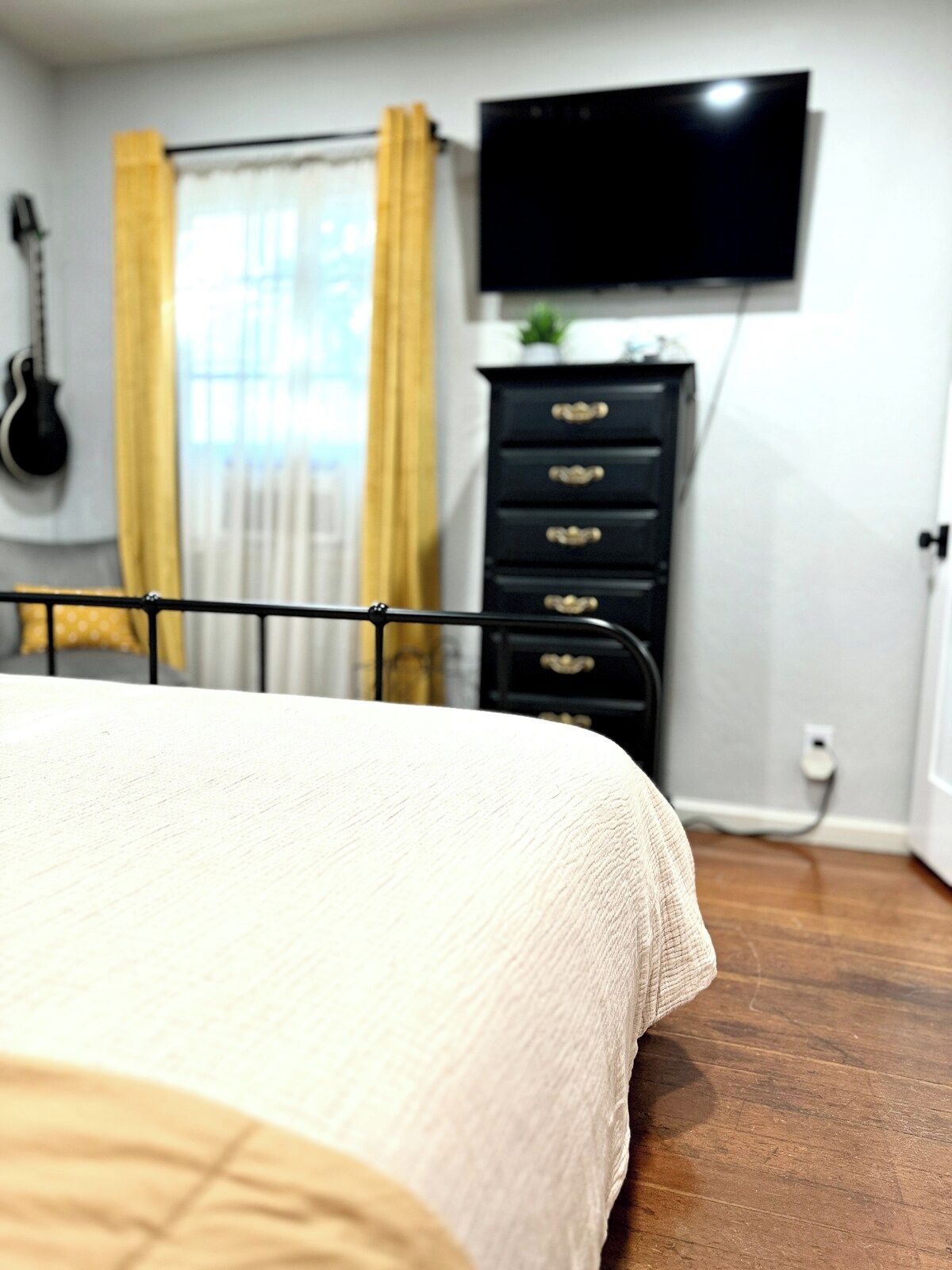 A bedroom with a bed, a dresser, a television mounted on the wall, and a guitar hanging on the wall.