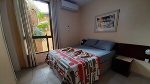 Una camera d'albergo con un letto, un comodino, una finestra con tende, un condizionatore d'aria e un quadro appeso al muro.