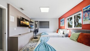 Une chambre d’hôtel équipée d’un lit, d’un bureau, d’un ventilateur, d’une télévision et décorée avec des œuvres d’art aux murs.