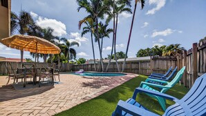 Un jardin avec une piscine, des chaises longues, une table avec des chaises et un parasol en chaume.