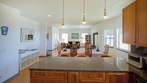 A spacious kitchen with granite countertops, wooden cabinets, and a central island. There are pendant lights, a dining area with a table and chairs, and a view of the outdoors through windows. -  Overlooking the ocean, sleeps 7, kid and pet friendly, pergola with privacy. (Whitethorn)