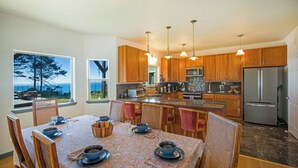 A modern kitchen with wooden cabinets, a stainless steel refrigerator, and a dining area with a table set for four. -  Overlooking the ocean, sleeps 7, kid and pet friendly, pergola with privacy. (Whitethorn)