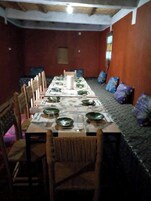 Una larga mesa de comedor dispuesta con platos, vasos y cubiertos, rodeada de sillas de madera en una habitación con paredes rojas y un suelo estampado.