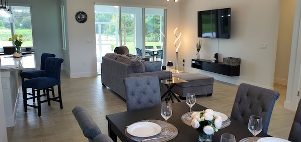 A modern living room with a sofa, dining table, and chairs. A TV is mounted on the wall, and there is a large window providing a view of the outdoors. - Waterski & More - Southwest facing pool (Clermont)