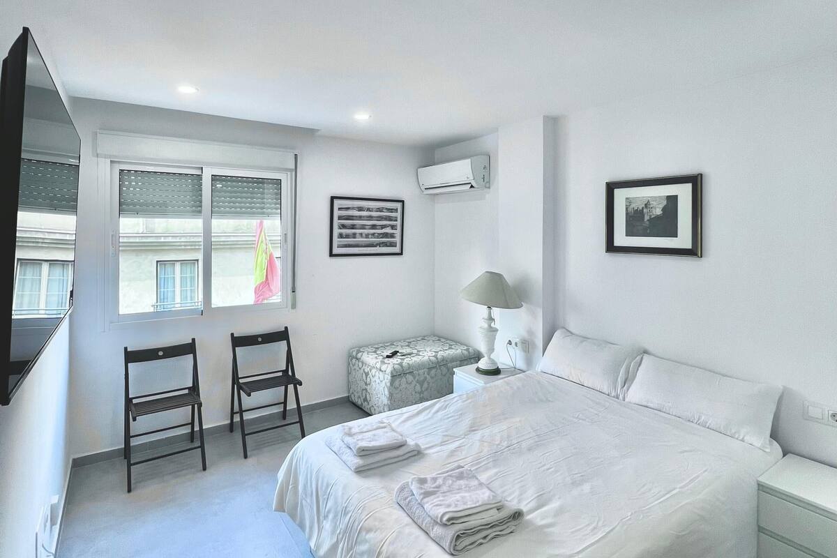 A bedroom with a large bed, a chair, a small table, and a window with blinds.