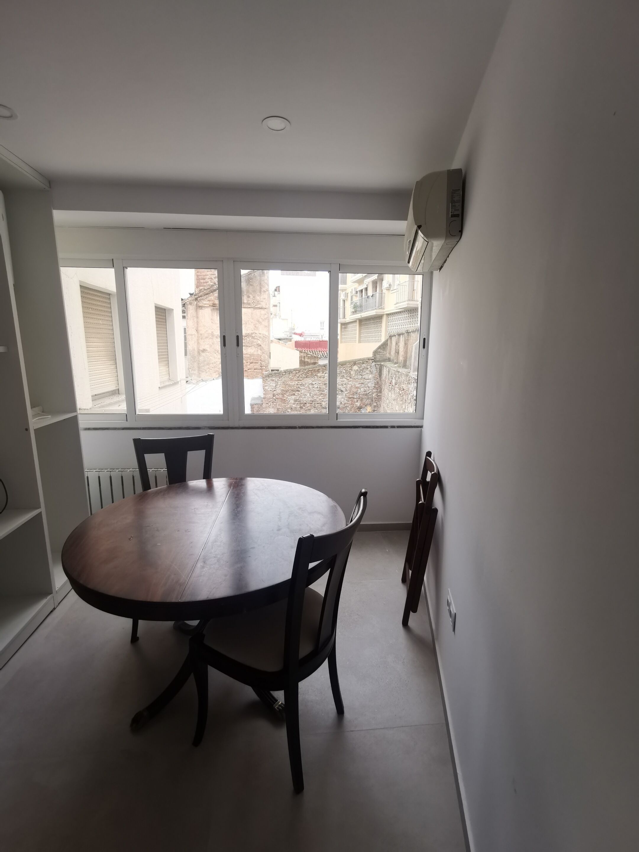 Un área de comedor con una mesa redonda y sillas, ventanas amplias con vistas a edificios, y un equipo de aire acondicionado en la pared.