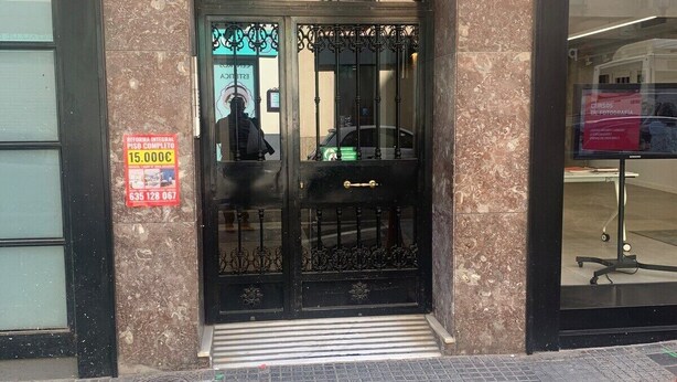 Entrada de un edificio con fachada de mármol, portón de metal negro y un cartel que indica un precio de habitación completo de 15.000€.