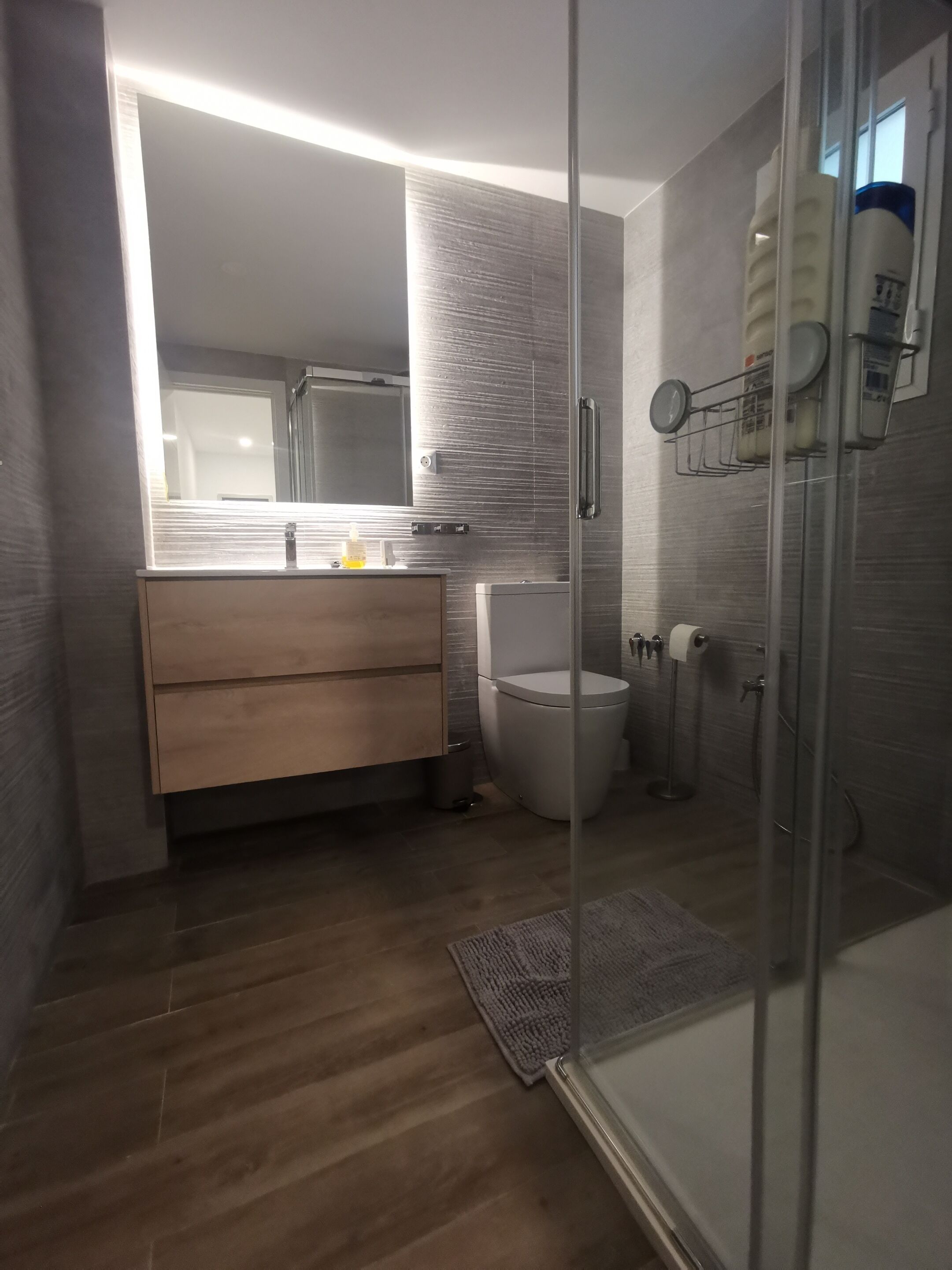 Een moderne badkamer met een douchecabine van glas, een houten wastafelmeubel, een toilet en een grote spiegel.
