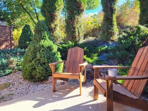 Dos sillas de madera tipo Adirondack y una mesita baja en un jardín con diversas plantas y una cerca de madera.