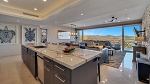 A modern kitchen with a large island, a dining area with a view, and a living space with a large window overlooking a desert landscape. - Luxury Cabo Condo in Pedregal, 2 bedroom, 2 bath, City & Ocean Views (Cabo San Lucas)