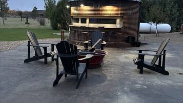 A rustic outdoor seating area with Adirondack chairs and a fire pit, surrounded by a wooden cabin with a metal roof.