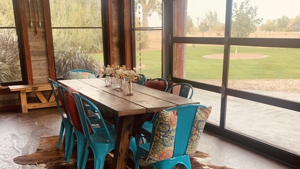 Ein rustikaler Essbereich mit Holztisch und türkisfarbenen Stühlen, einem Kuhfellteppich und großen Fenstern mit Blick auf eine Wiese.