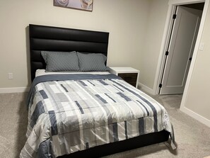 Un lit bien fait, recouvert d’une couvre-lit à motifs, avec une tête de lit noire et deux oreillers. Une table de chevet blanche, dotée d’un tiroir, se trouve à côté du lit. Une porte est visible sur la droite.
