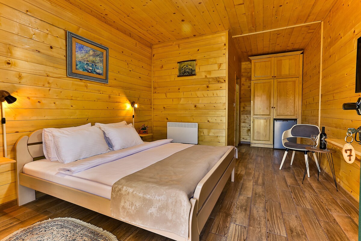 A wooden cabin room with a bed, a chair, a table with a bottle and glasses, and a door leading to another room.