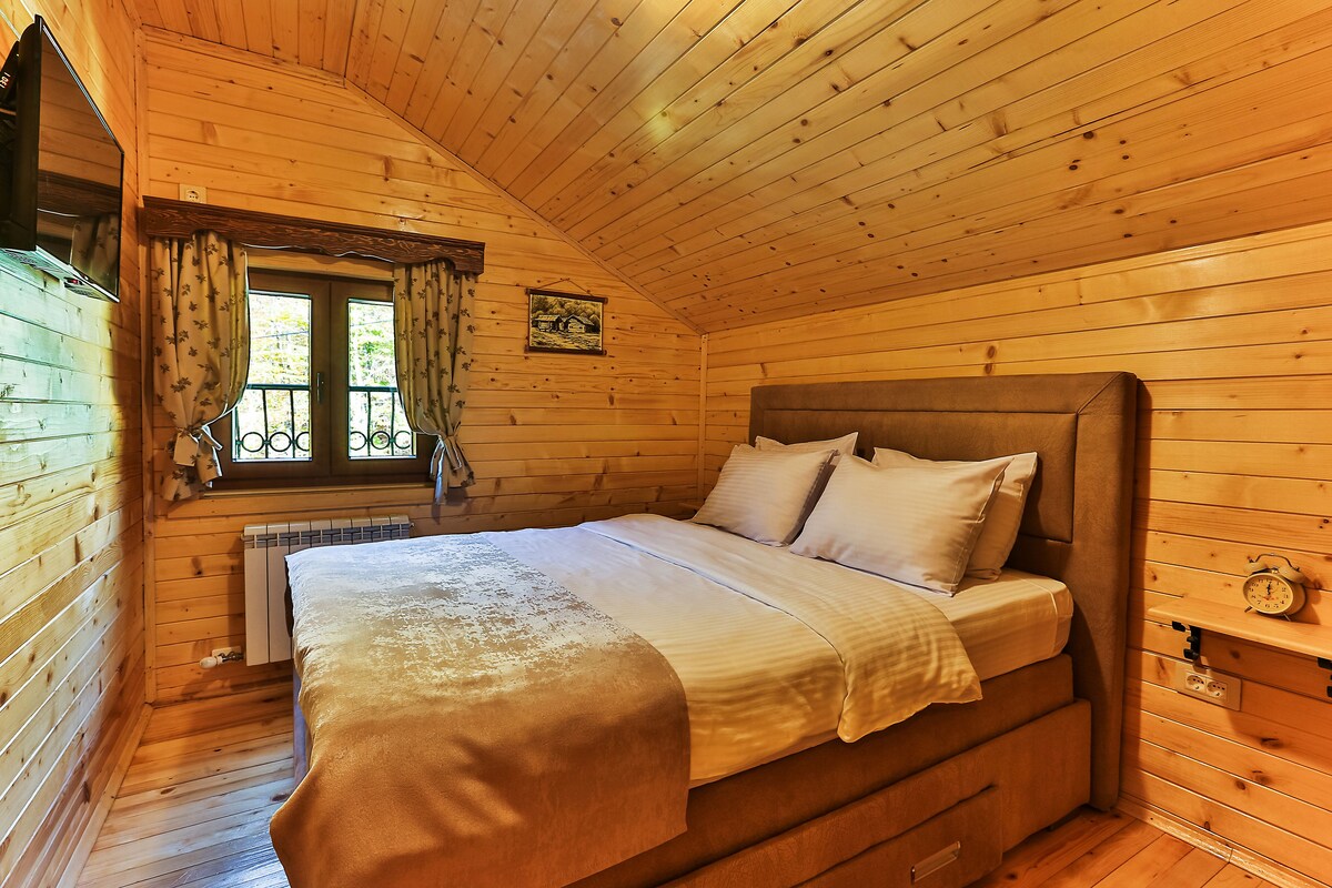 A wooden cabin bedroom with a bed, a window with curtains, a radiator, and a picture on the wall.
