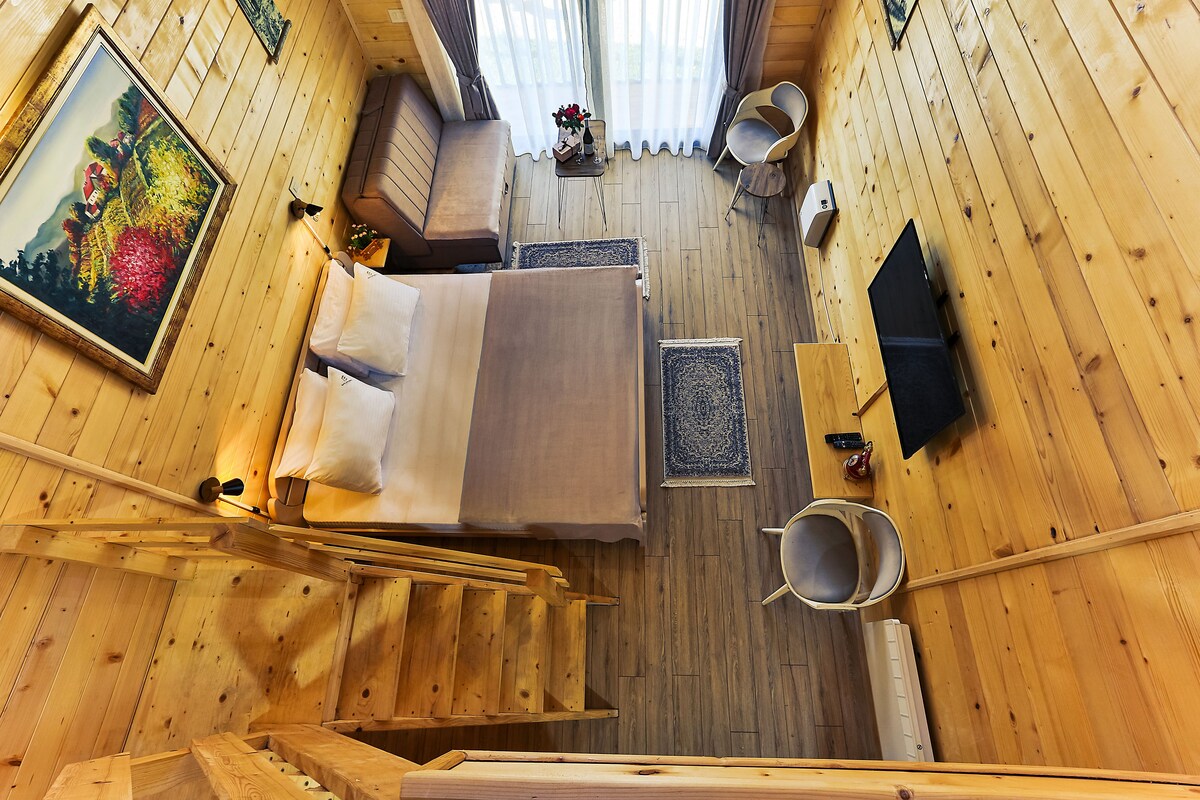 A wooden cabin with a bed, a small table with a vase, a chair, and a painting on the wall.
