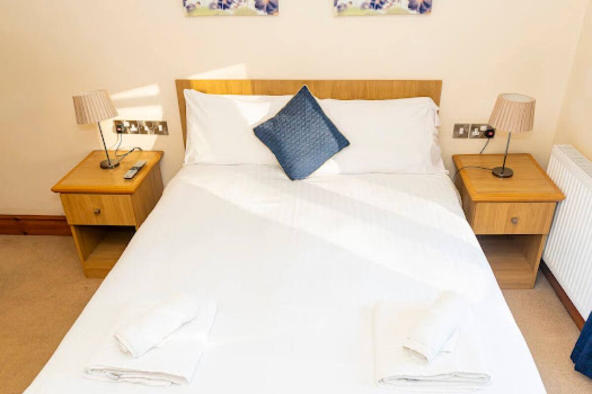 A neatly made bed with white linens, a blue pillow, and two folded towels on the bed. There are two wooden nightstands with lamps and a power outlet on each.