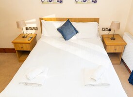 Una cama bien hecha con sábanas blancas, una almohada azul y dos toallas dobladas sobre la cama. Hay dos mesitas de noche de madera, cada una con una lámpara y un enchufe.