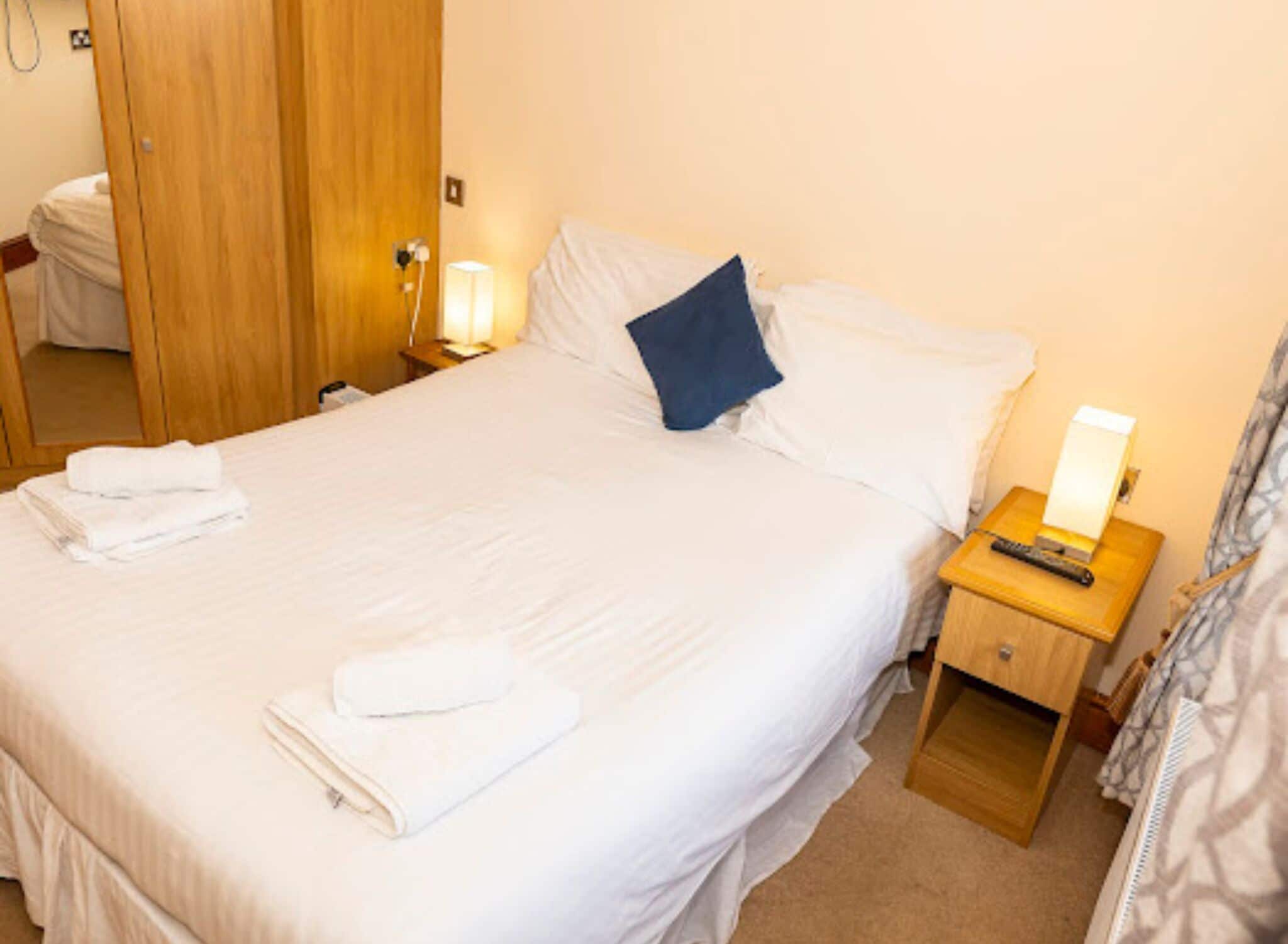 A neatly made bed with white linens and a blue pillow, a bedside lamp, and a wooden nightstand with a remote control.