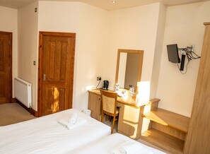 Ein Hotelzimmer mit zwei Betten, einem Holztisch mit Spiegel, einem Heizkörper und einem an der Wand montierten Fernseher.