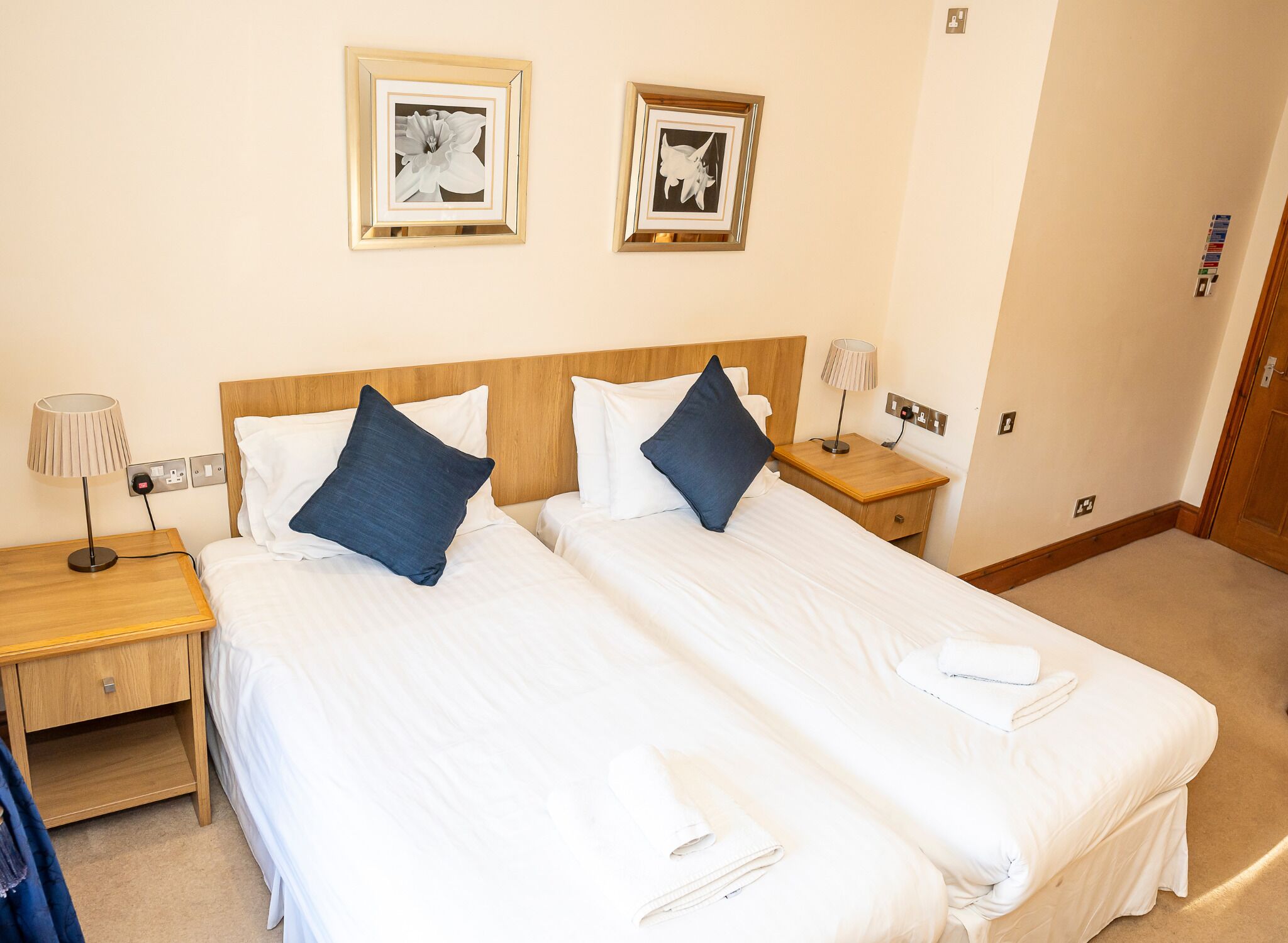 A hotel room with two beds, each with white linens and blue pillows. There are two framed pictures on the wall, a nightstand with a lamp, and a wooden headboard.