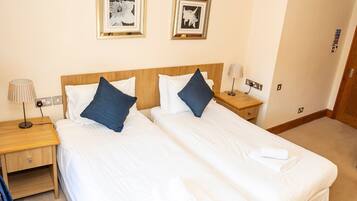 A hotel room with two beds, each with white linens and blue pillows. There are two framed pictures on the wall, a nightstand with a lamp, and a wooden headboard.