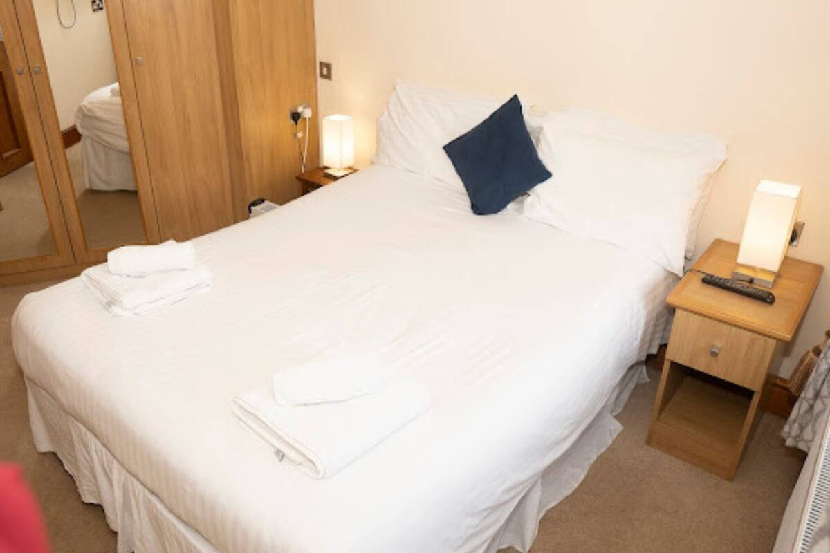 A neatly made bed with white linens and a folded towel, a bedside lamp, and a wooden nightstand with a remote control.