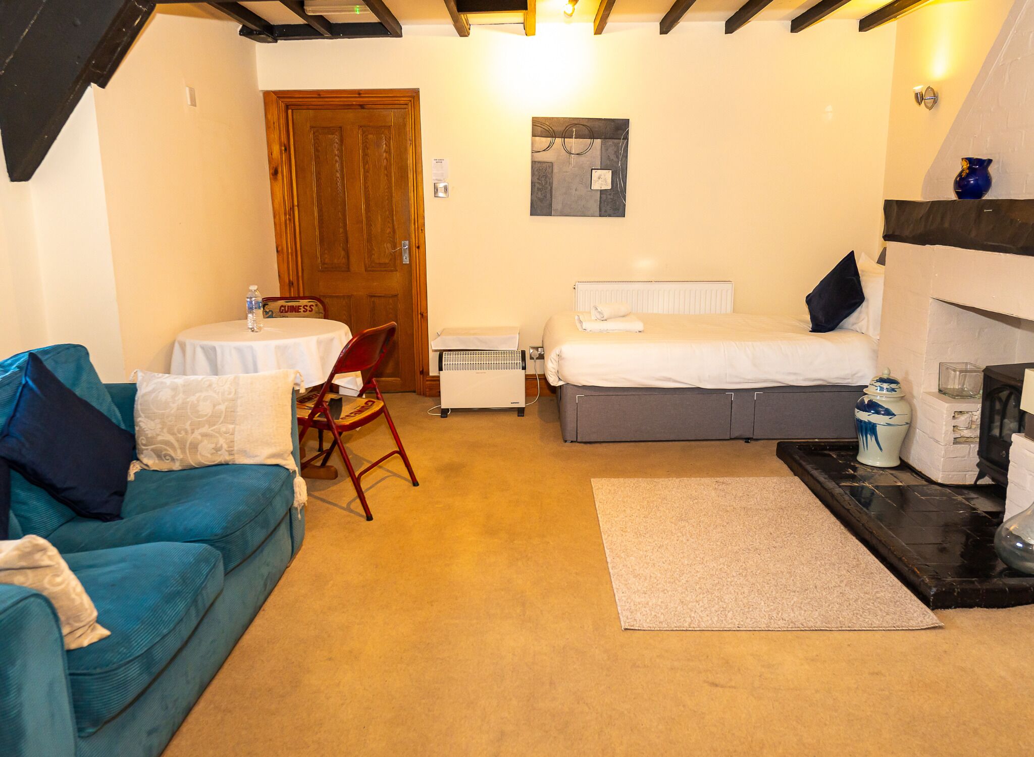 A compact room with a blue sofa, a small table with a chair, a bed with a white mattress, and a radiator.