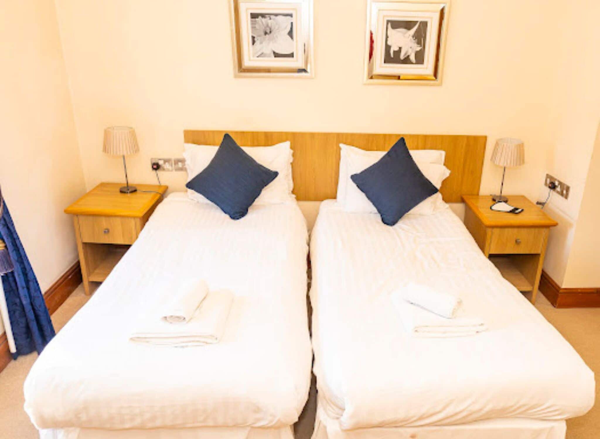 A hotel room with two single beds, each with a pillow and a folded towel. There are bedside tables with lamps and a wall-mounted phone.