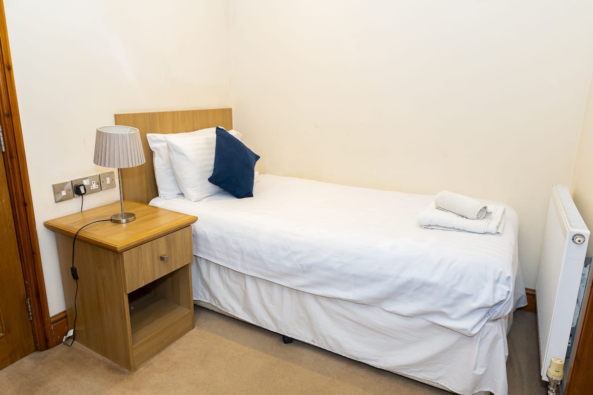 A neatly made bed with white bedding and a blue pillow, a bedside table with a lamp, and a power outlet.