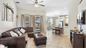 A spacious living room with a brown leather sofa, a coffee table, and a ceiling fan. A kitchen area with a bar and a dining table is visible in the background. - The Minnion's Palace Villa (Kissimmee)