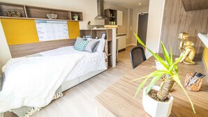 Moderni hotellihuone, jossa on sänky, työpöytä, tuoli, pieni keittotila ja ruukkukasvi.