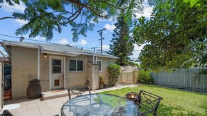 A house with a patio area featuring a glass table and chairs, a fire pit, and a small garden with potted plants and a fence.
