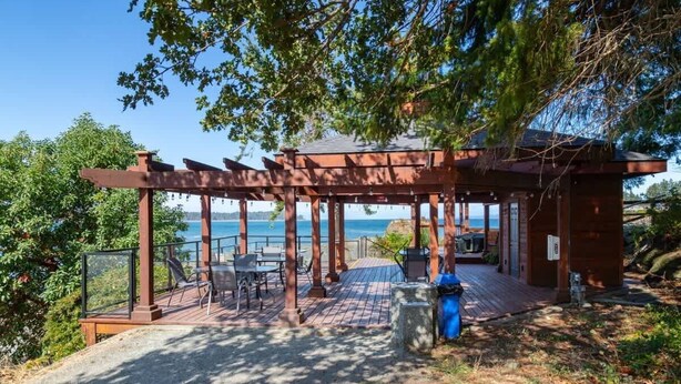 A wooden pergola with a view of the sea, outdoor seating, and a small building. - Serenity on the Bay - Studio (Nanoose Bay)