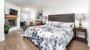 A bedroom with a large bed, a fireplace, a sofa, and a TV mounted on the wall. - Serenity on the Bay - Studio (Nanoose Bay)