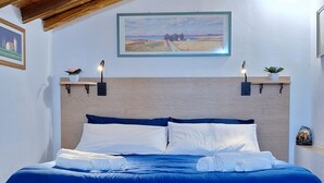Un dormitorio con techo de madera, una cama con ropa de cama azul, cabecera de madera, alfombra estampada con motivos florales y dos lámparas fijadas a la pared.