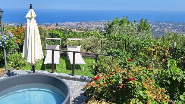 Zona de piscina con vista al mar, rodeada de abundante vegetación y una pérgola con sombrilla blanca.