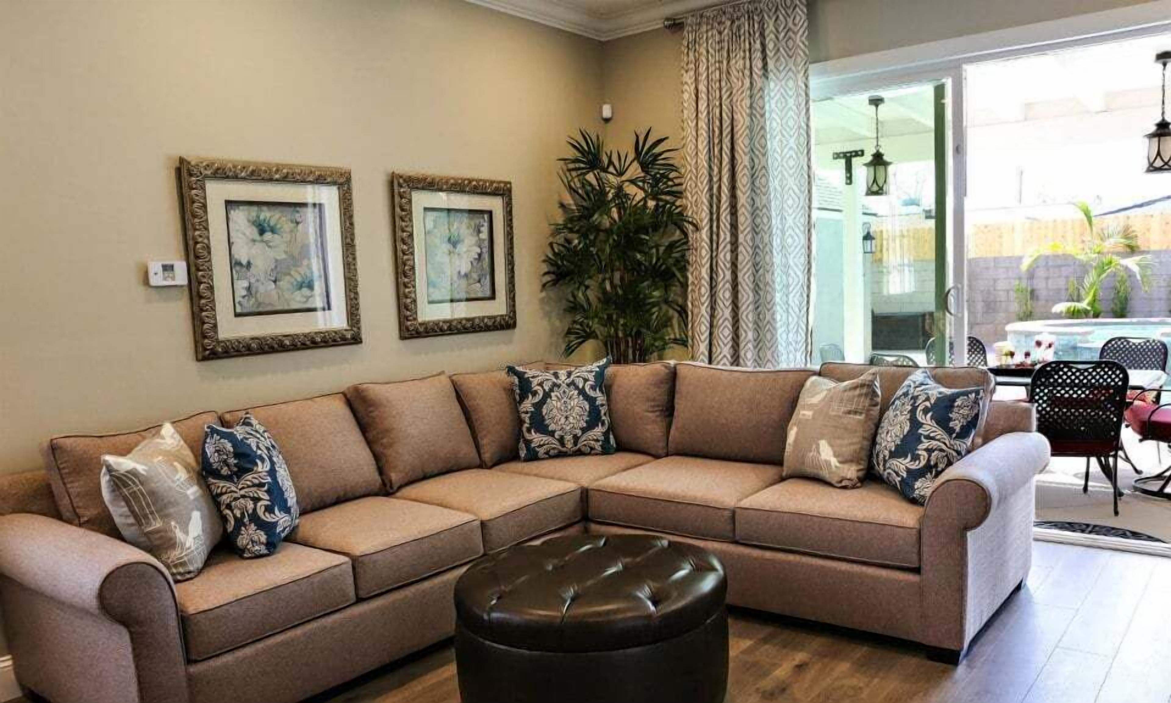 A living room with a beige sectional sofa, two framed artworks, a potted plant, and a view of an outdoor patio area.