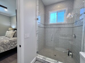 A bathroom with a shower, a window, and a bed in the adjacent room.