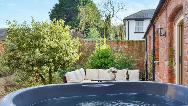 Un jacuzzi en un jardín con un muro de ladrillo, una casa blanca y varios árboles.
