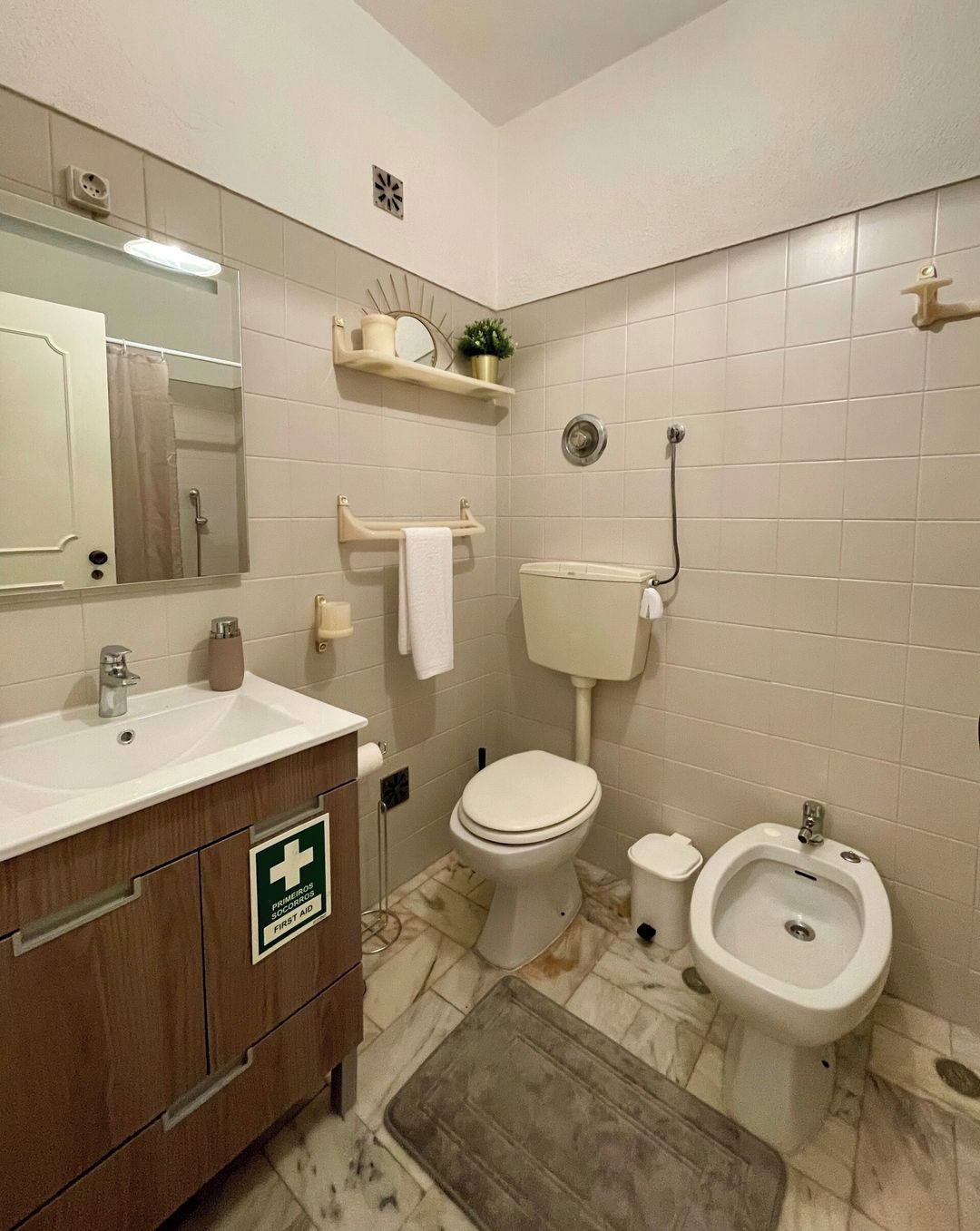 A bathroom with a toilet, bidet, sink, and a first aid sign on the cabinet.