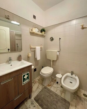 Casa de banho com sanita, bidé, lavatório e um sinal de primeiros socorros na gaveta.