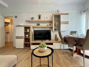 Un soggiorno moderno con TV a schermo piatto, mensole in legno, un'area pranzo con tavolo imbandito per due e un piccolo tavolino con una pianta.