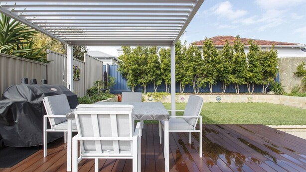 Un área de patio techada con piso de madera, pérgola de metal blanco y mobiliario exterior.