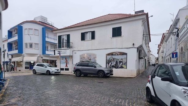 Uma rua de paralelepípedos com edifícios, incluindo um prédio branco com uma placa que diz 'BILLA BONG'.