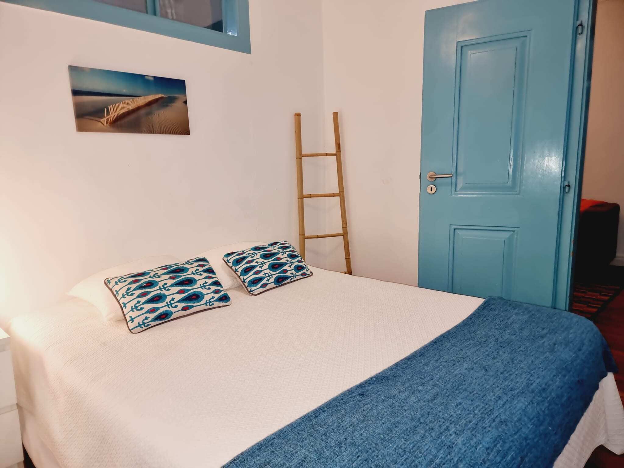 Un lit avec un drap blanc et une couverture bleue, deux coussins à motifs, une échelle en bois et une porte bleue.