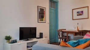 Uma sala de estar com televisão de tela plana, um rack branco, uma porta azul, uma mesa de jantar com cadeiras e um tapete colorido.