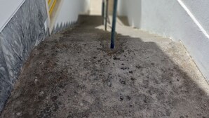 Un étroit chemin de béton mène à l’entrée d’un bâtiment, qui est agrémentée d’une rambarde métallique et d’un panneau de ventilation jaune.