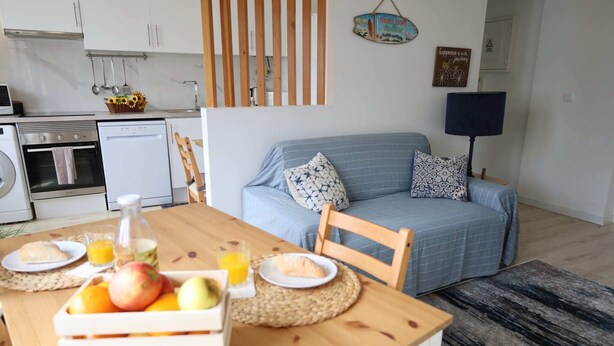 A modern kitchen with white cabinets, a dining table with a fruit basket, and a blue sofa with patterned cushions. - Mocho's  - Apartment (Mafra)