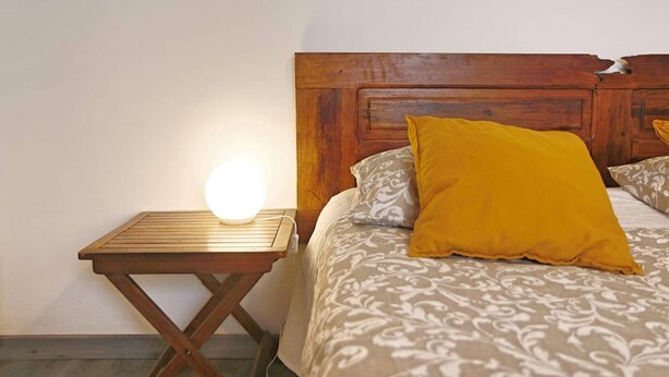 Una cama con una colcha estampada y una almohada amarilla, una mesita de noche de madera con una lámpara y un cabecero de madera.